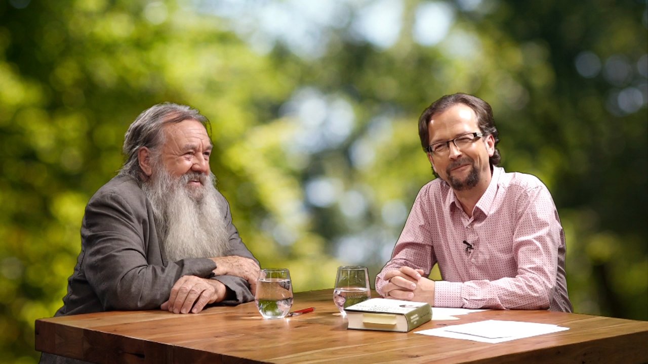 Wolf-Dieter Storl sitzt neben Moderator Thomas an einem Tisch. Thomas blickt in die Kamera.