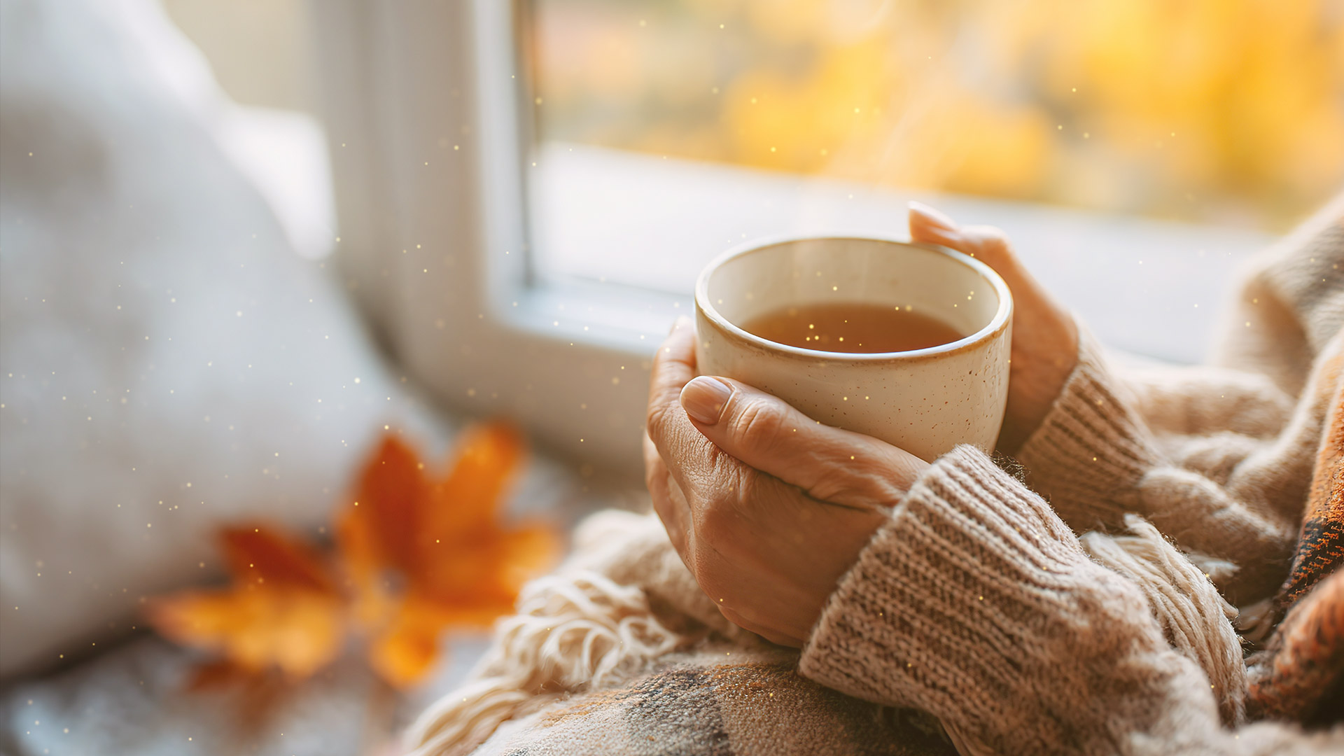 In ein weiches Plaid gehüllte Hände halten eine dampfende Tasse Tee, im Hintergrund goldene Herbstblätter. Die Szene ist in warmes Licht getaucht. 