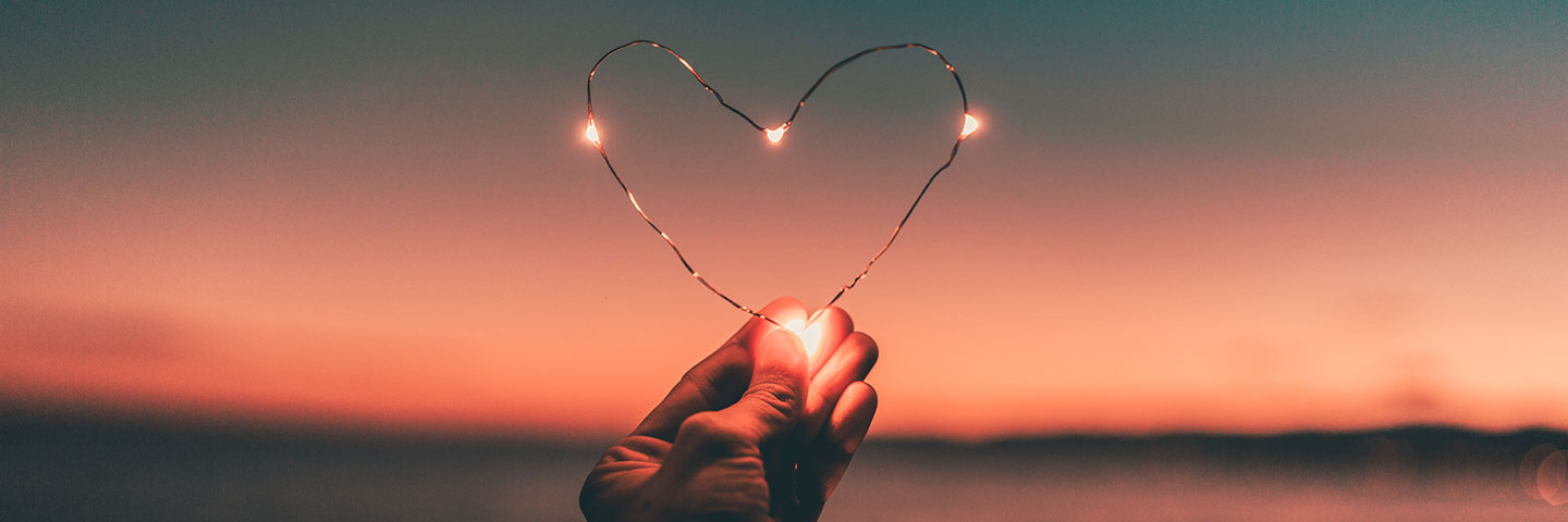 Vor einem Sonnenuntergang hält eine Hand ein Herz aus Draht in die Kamera. Das Herz leuchtet an mehreren Stellen.