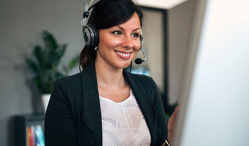 Eine Frau trägt ein Headset und schaut lächelnd auf einen Monitor.