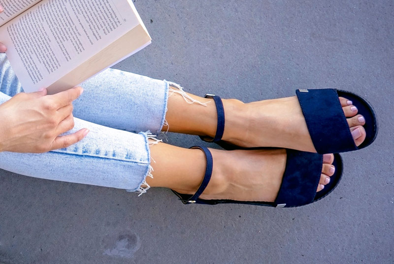 Blick von oben auf die Beine einer Frau, die dunkelblaue Sandalen zu hellblauen Jeans trägt und ein Buch in der Hand hält.