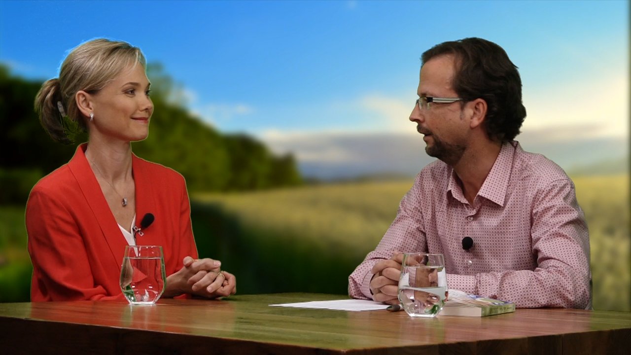 Jana Haas sitzt mit dem Moderator Thomas an einem Tisch. Sie sehen einander an und lächeln.