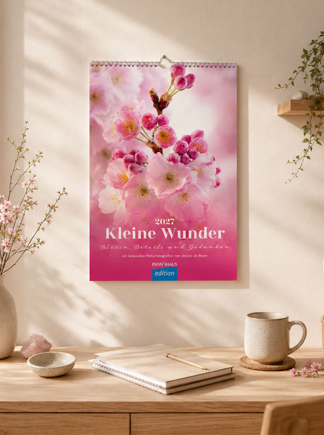 Wandkalender „Kleine Wunder 2027“ mit rosa Kirschblüten-Motiv an einer hellen Wand über einem Schreibtisch. Umgeben von Vase mit Blütenzweigen, Tasse, Notizbuch und einem Wandregal mit Pflanzen.