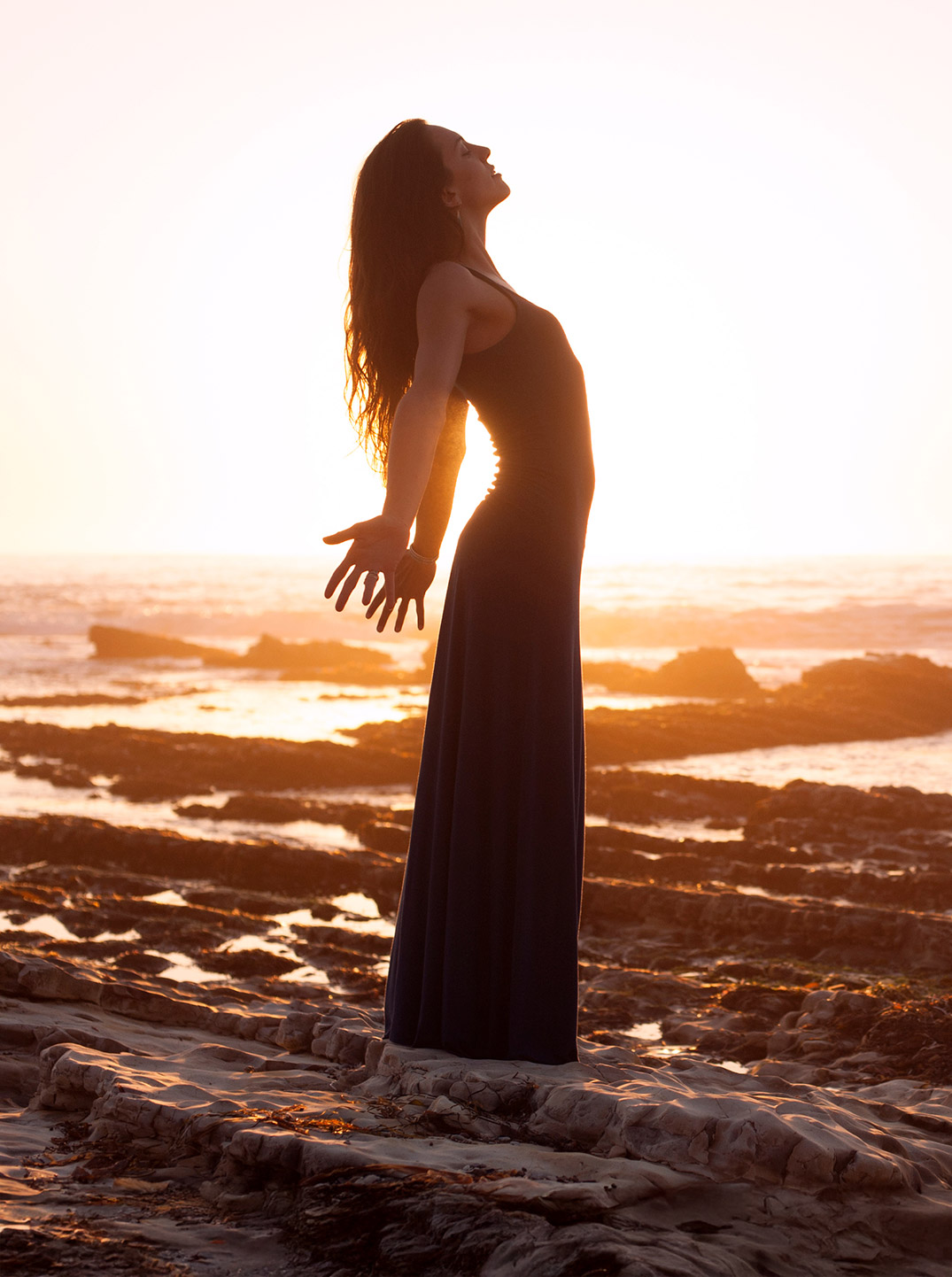 Frau im langen Kleid steht barfuß auf Felsen am Meer, breitet die Arme aus und genießt im warmen Sonnenuntergang ein Gefühl von Freiheit und Weite.