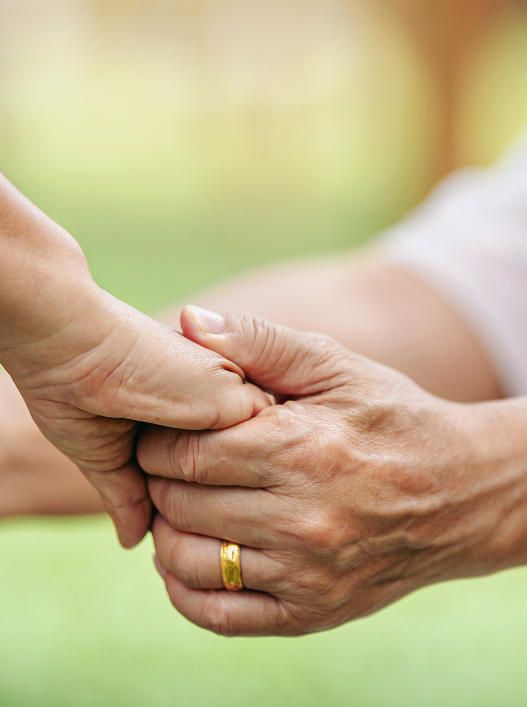 Eine ältere Hand hält behutsam die Hand einer anderen Person vor einem weich verschwommenen grünen Hintergrund.