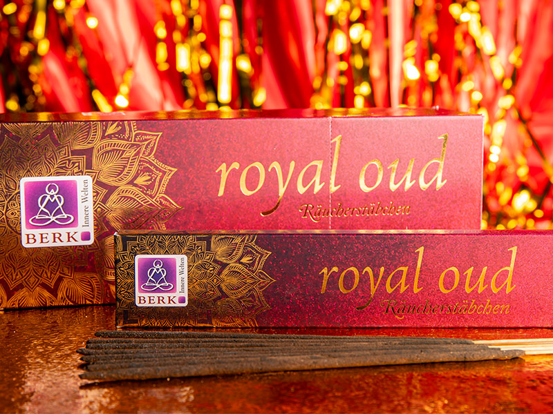 Mehrere Packungen Räucherstäbchen der Sorte „Royal Oud“ liegen auf einer glitzernden rot-goldenen Oberfläche. Davor sind mehrere dunkle Räucherstäbchen ausgebreitet.