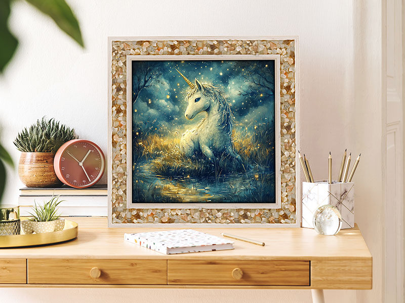 Ein Energiebild zeigt ein weißes Einhorn, das an einem Wasserlauf inmitten einer märchenhaften Waldlichtung ruht. Der Rahmen ist mit Mondstein besetzt. Das Bild steht auf einem hellen Schreibtisch, angelehnt an die Wand.