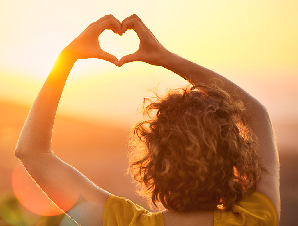 Person blickt in den Sonnenuntergang und formt mit den Händen ein Herz vor der tief stehenden Sonne. Warmes goldenes Licht, sanfte Hügel und weiche Lichtreflexe vermitteln ein Gefühl von Lebensfreude, Dankbarkeit und Verbundenheit.