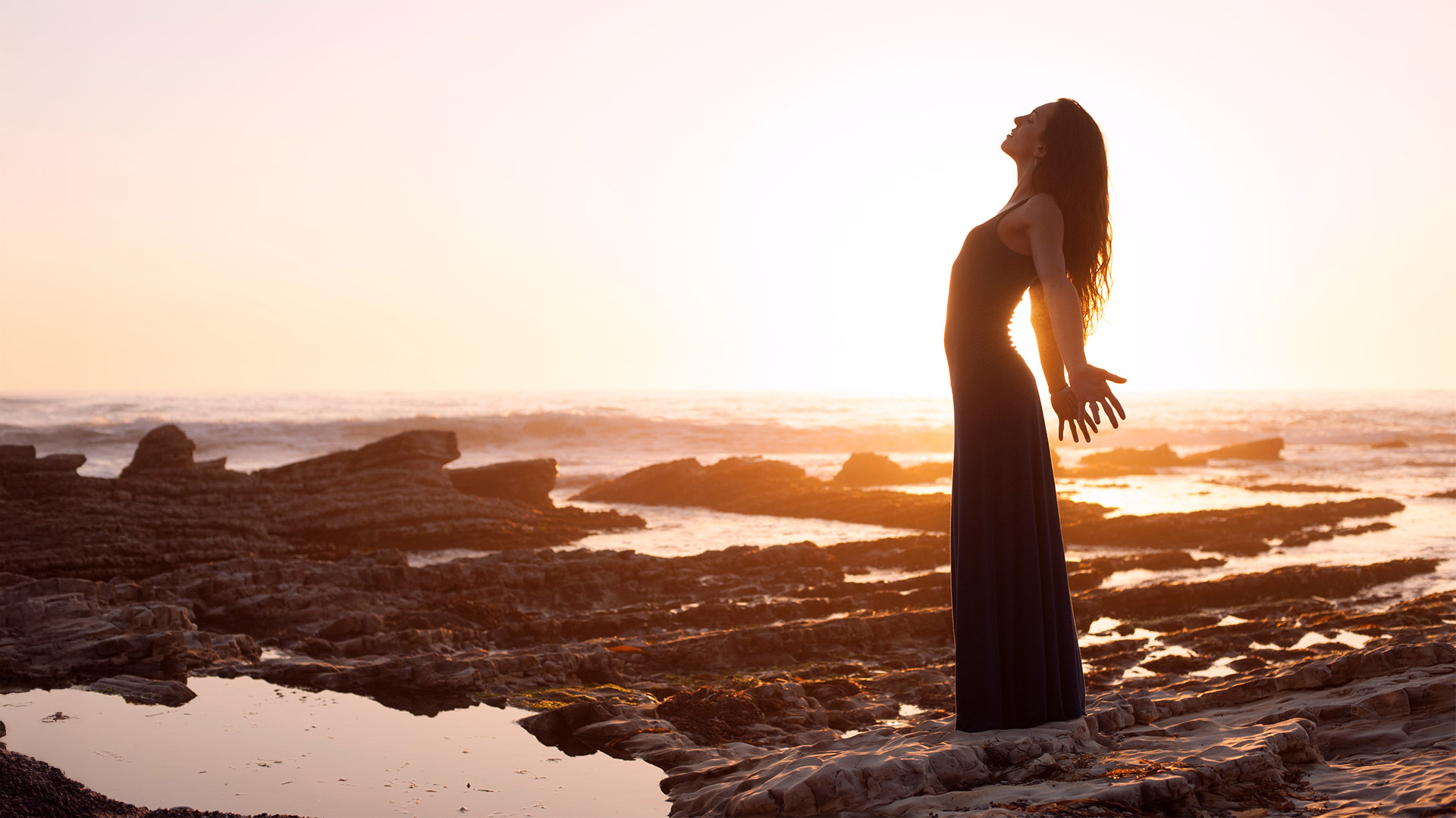 Frau im langen Kleid steht barfuß auf Felsen am Meer, breitet die Arme aus und genießt im warmen Sonnenuntergang ein Gefühl von Freiheit und Weite.