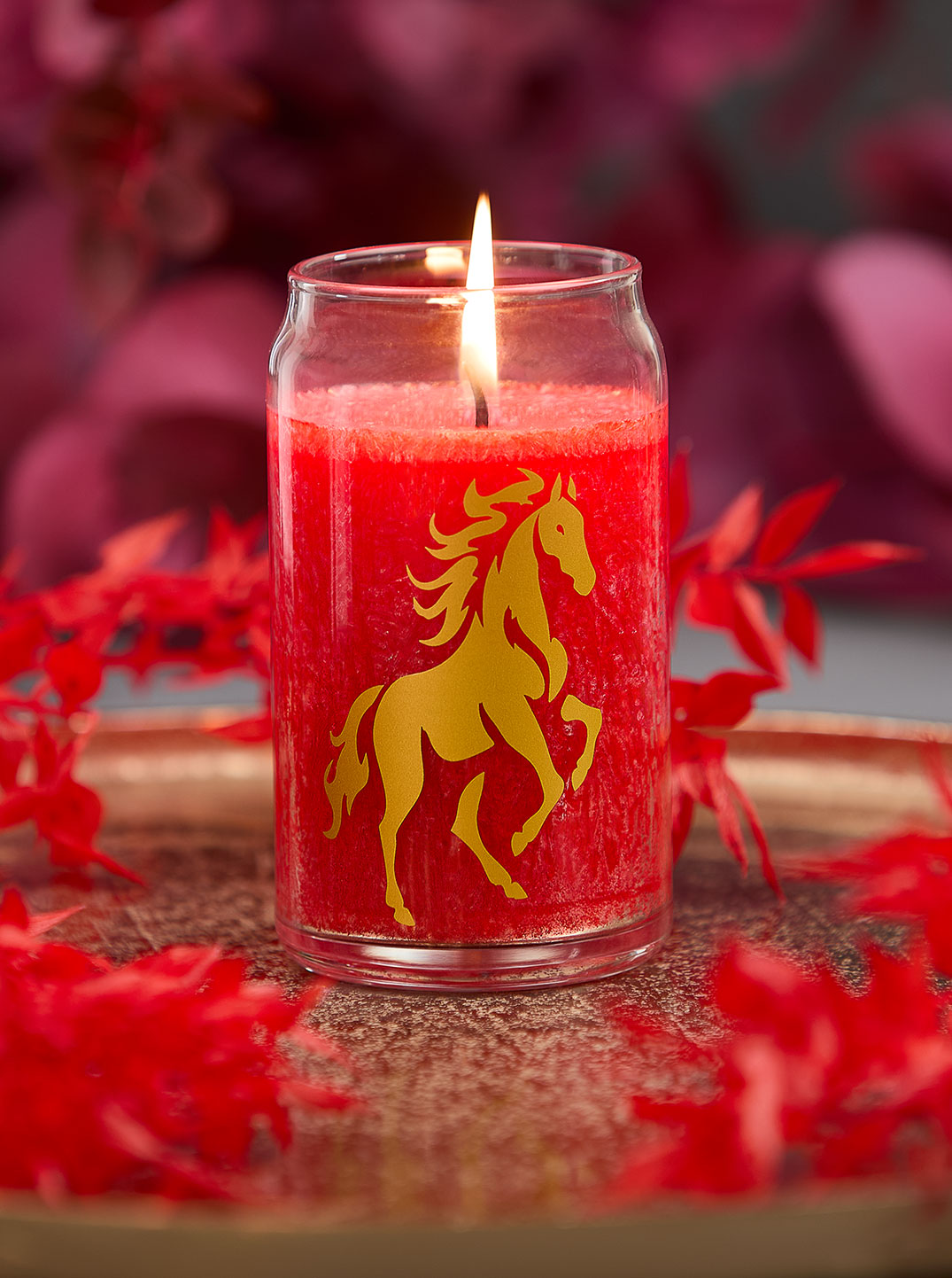 Leuchtend rote Kerze im Glas mit goldenem Pferdemotiv steht auf einem Kupferteller, umrahmt von roten Blättern – ein ausdrucksstarkes Arrangement voller Kraft, Eleganz und spiritueller Symbolik für das Jahr des Feuerpferds.