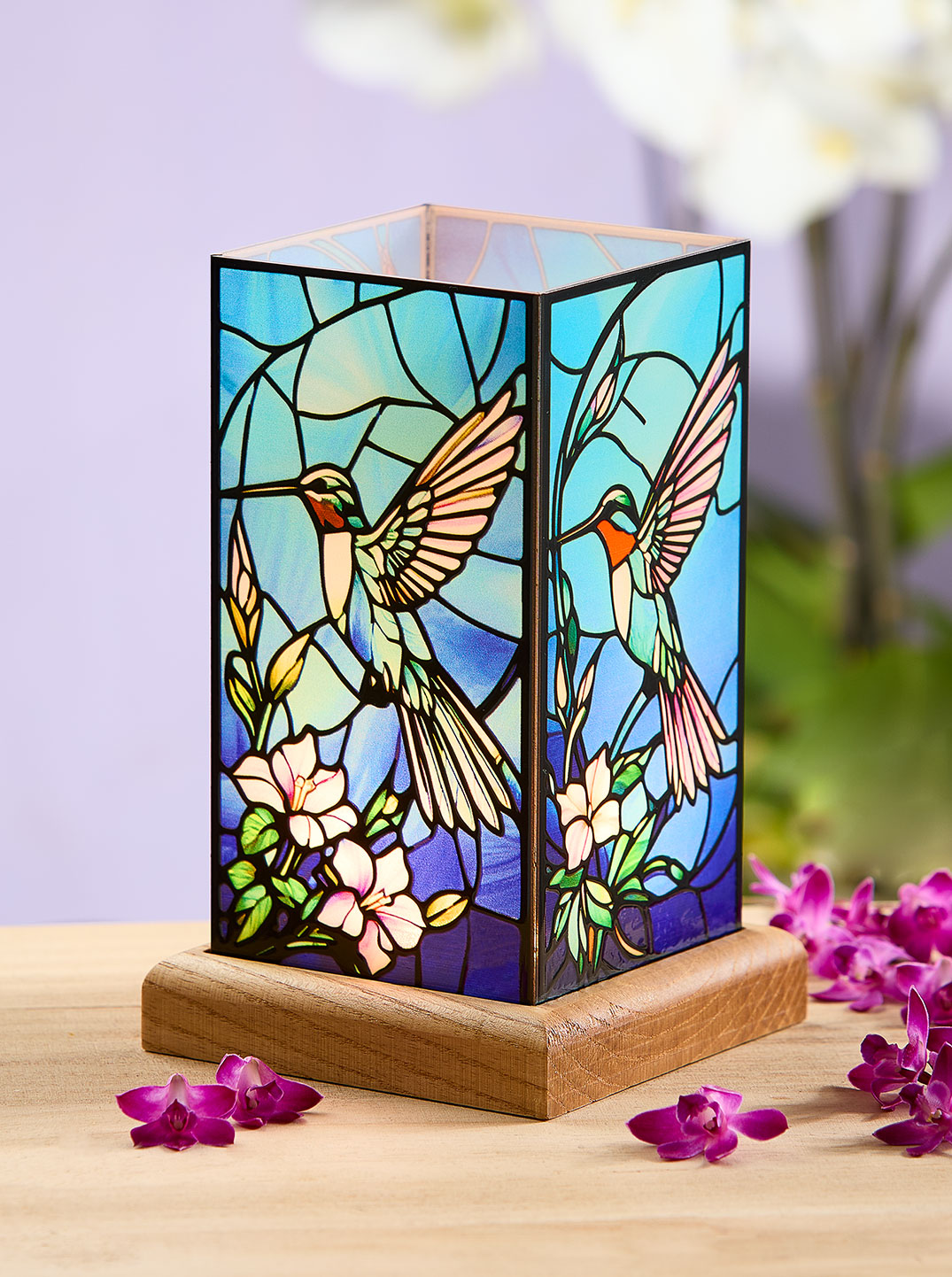 Quadratische Tischlampe im Tiffany-Stil mit farbigem Glasmotiv aus Kolibris und Blüten auf Holzsockel. Daneben liegen violette Blüten, links ein Hinweis „-20 %“. Die Szene wirkt dekorativ, lichtvoll und bringt Farbe ins zu Hause.