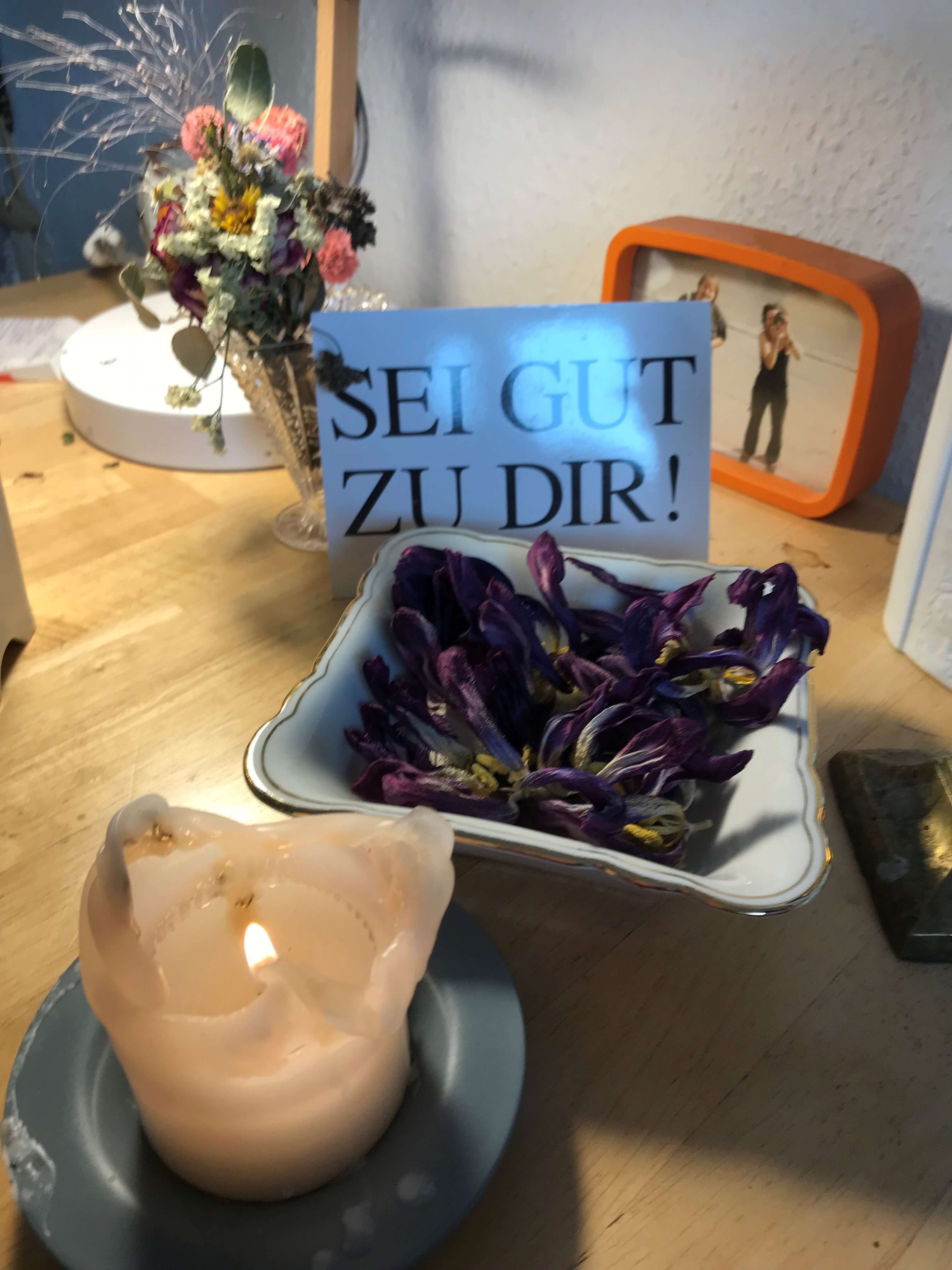 Brennende Kerze neben getrockneten Blüten und einer Postkarte mit den Worten "Sei gut zu dir!"