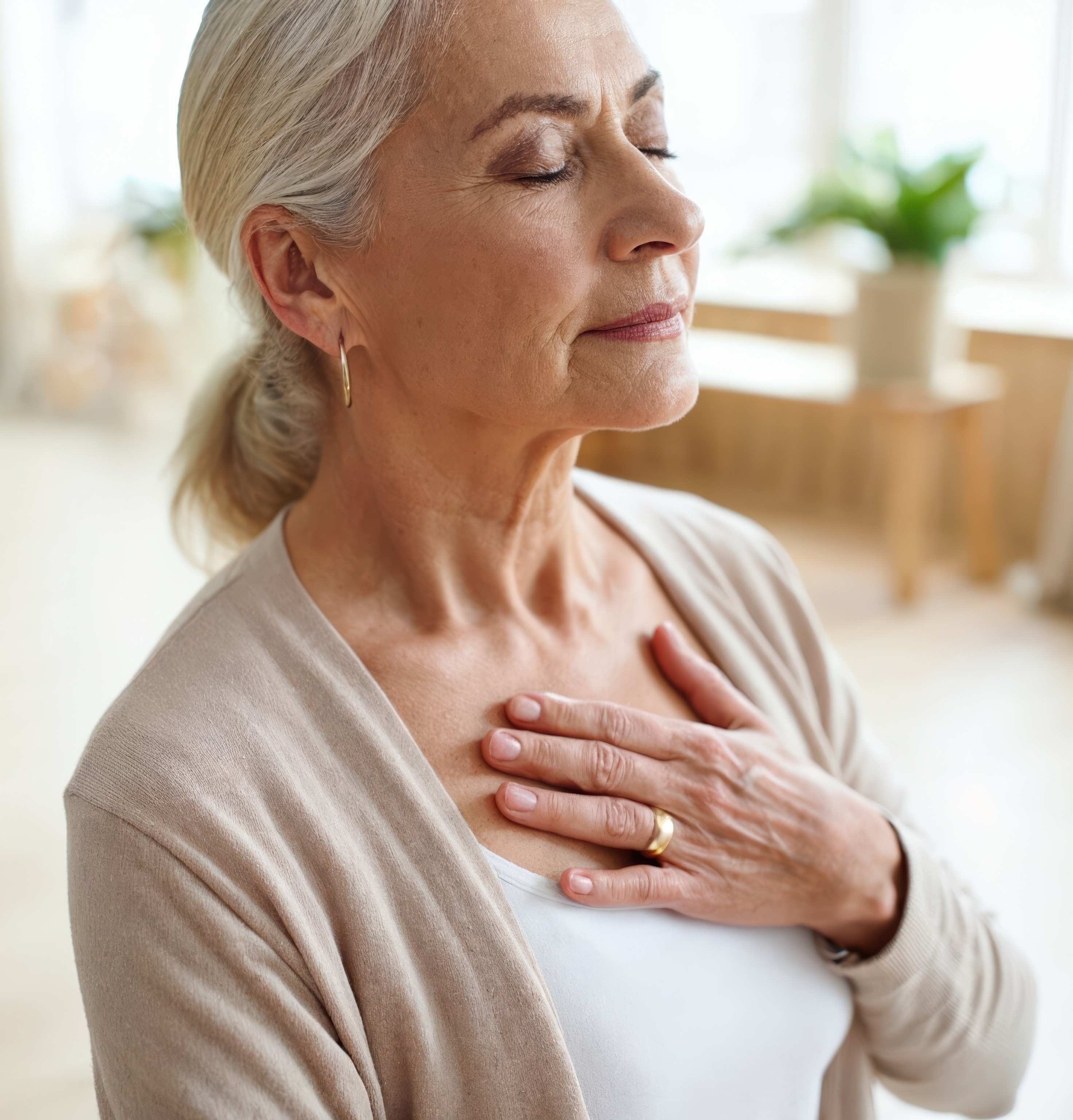 Ältere Frau mit geschlossenen Augen legt in hellem Raum ruhig eine Hand auf die Brust.